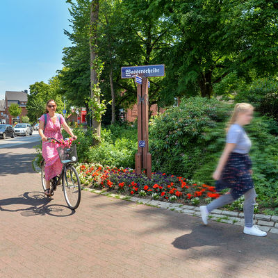 Bild der Wasserscheide, davor zwei Personen auf Fahrrädern und eine Fußgängerin, im Hintergrund mehrere Fahrzeuge in der Rathausstraße.
